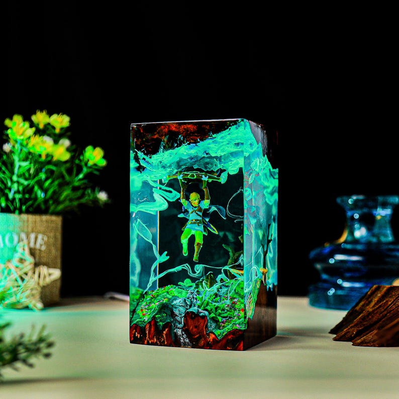May include: A rectangular resin sculpture featuring a miniature figure suspended within a glowing, teal-colored environment. The sculpture has a dark base with red and green accents, and the figure is holding a weapon. The overall theme is fantasy.