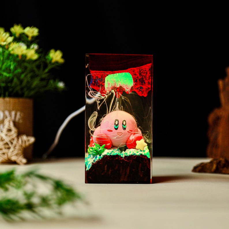 May include: A rectangular resin night light featuring a pink cartoon character with large green eyes. The light has a red and green glow, with white wisps throughout. The base is dark brown, and the light is plugged in with a white cord.