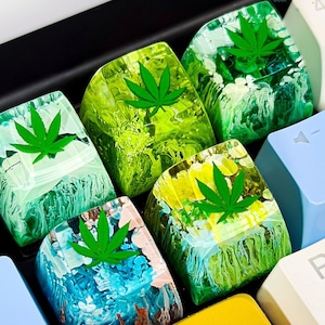 May include: Six green and white resin keycaps with a green marijuana leaf design. The keycaps are in a variety of swirling patterns.