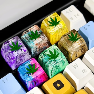 May include: A set of 8 colorful keycaps featuring a green marijuana leaf design. The keycaps are made of resin and have a unique marbled effect. The keycaps are designed to fit standard mechanical keyboards.