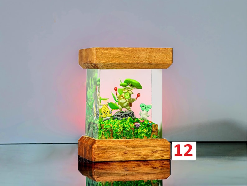 K&ouml;nnte beinhalten: Eine Holzkiste mit einer Glasfront, die ein Miniatur-Terrarium mit gr&uuml;nen Pflanzen, einer kleinen wei&szlig;en Figur und einer roten Beere enth&auml;lt. Die Kiste ist von innen beleuchtet und erzeugt ein warmes Leuchten auf die Szene. Die Zahl "12" ist auf einem wei&szlig;en Etikett geschrieben.