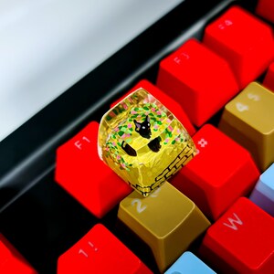 K&ouml;nnte beinhalten: Eine gelbe und gr&uuml;ne Resin-Tastenkappe mit einem schwarzen Katzenmotiv. Die Tastenkappe liegt auf einer roten mechanischen Tastatur.