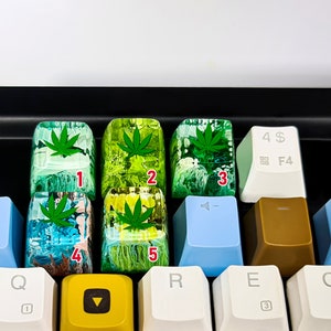 May include: Five green and yellow resin keycaps with a marijuana leaf design. The keycaps are numbered 1 through 5.