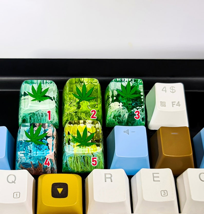 May include: Five green and yellow resin keycaps with a marijuana leaf design. The keycaps are numbered 1 through 5.