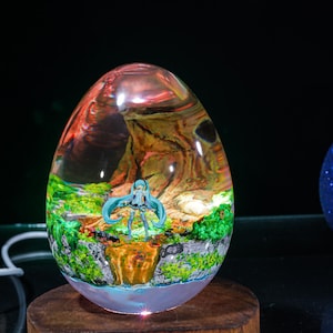 Puede incluir: Un pisapapeles de resina transparente en forma de huevo que contiene una figura en miniatura. La figura es un personaje de anime con cabello azul de pie en una escena de bosque en miniatura. La resina es transparente y muestra los detalles de la figura y la escena del bosque.