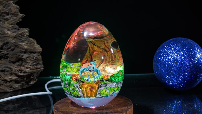 Puede incluir: Un pisapapeles de resina transparente en forma de huevo que contiene una figura en miniatura. La figura es un personaje de anime con cabello azul de pie en una escena de bosque en miniatura. La resina es transparente y muestra los detalles de la figura y la escena del bosque.