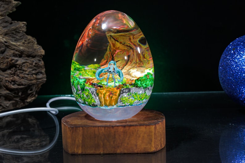 Puede incluir: Una base de l&aacute;mpara de vidrio transparente en forma de huevo con una escena de resina colorida en su interior. La escena presenta un paisaje verde y marr&oacute;n con una figura de pelo azul de pie en el centro. La base est&aacute; sobre un soporte de madera.
