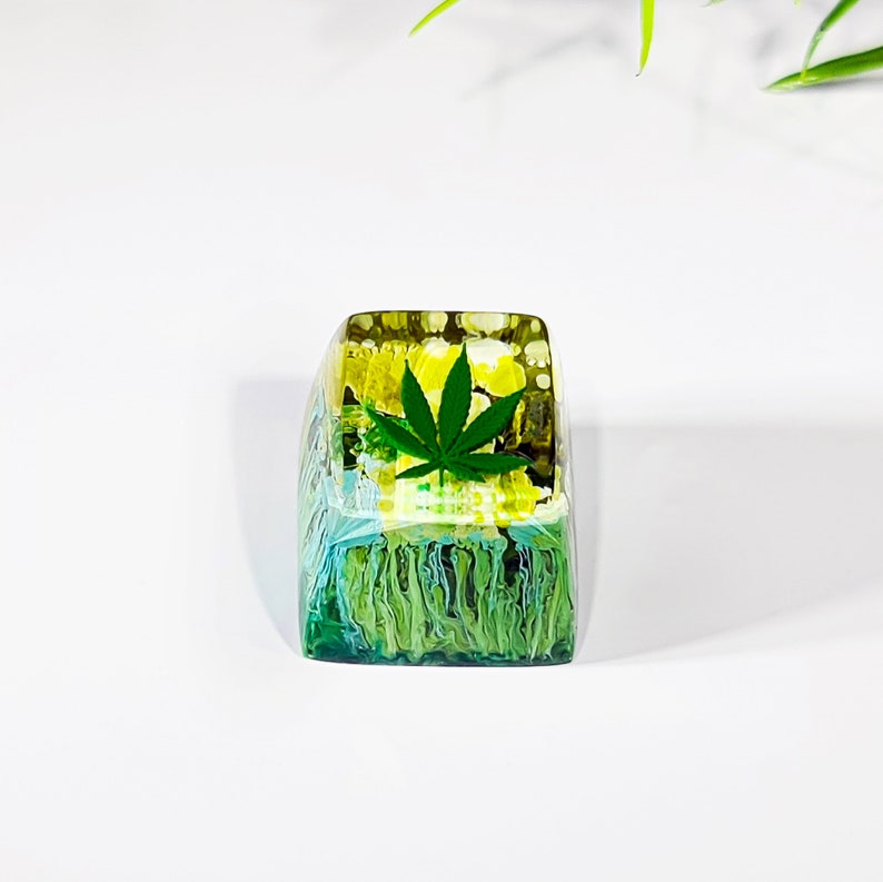 May include: A green marijuana leaf encased in clear resin with a yellow and green swirl pattern. The resin is shaped like a keyboard key.