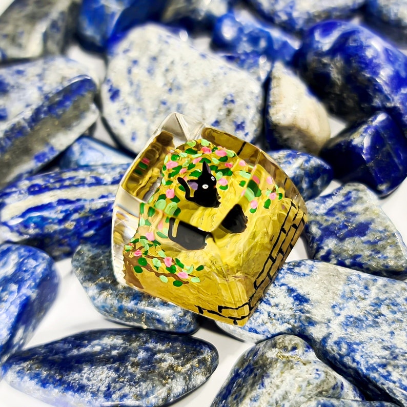 K&ouml;nnte beinhalten: Ein transparenter Keycap aus Kunstharz mit handbemaltem Design eines schwarzen Katers, der in einem gelben Backsteinhaus mit gr&uuml;nen Ranken und rosa Blumen sitzt.