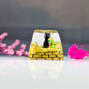 K&ouml;nnte beinhalten: Ein transparenter Harz-Tastenkappen mit einer schwarzen Katzenfigur, die auf einer gelben Backsteinmauer mit gr&uuml;nen und rosa Blumen sitzt.