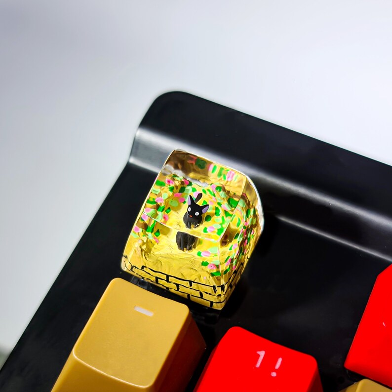 K&ouml;nnte beinhalten: Ein schwarzer Katzenfigur in einem klaren Harz-Tastenkappen mit einem gelben, gr&uuml;nen und rosa Blumendesign. Die Tastenkappe befindet sich auf einer schwarzen Tastatur.