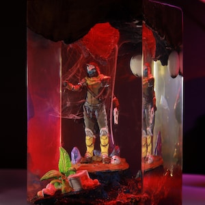 May include: A rectangular resin block containing a miniature figure of a humanoid character with a blue mask, yellow leg armor, and a weapon. The scene features red smoke effects, small white spheres, a green plant, and a skull on a rocky base.