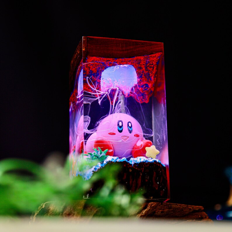May include: A rectangular resin art piece featuring a pink cartoon character with blue eyes, red feet, and a star. The character is surrounded by a jellyfish and other sea elements, encased in a clear resin block with a dark wood top.