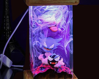 May include: A wooden lamp base with a clear resin insert containing a purple and black Pokemon character. The resin is filled with white swirls. The lamp is lit up with a purple glow. The lamp has a number "1" on the base.