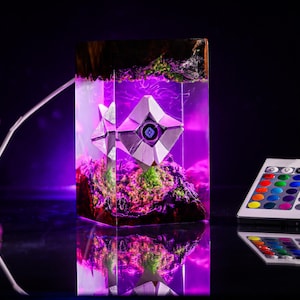 May include: A rectangular resin lamp featuring a white geometric Ghost character encased within clear resin, illuminated by a vibrant purple LED light. The base and top contain dark wood and green moss textures. Includes a multi-colored remote control.