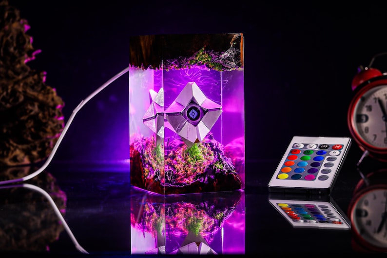 May include: A rectangular resin lamp featuring a white geometric Ghost character encased within clear resin, illuminated by a vibrant purple LED light. The base and top contain dark wood and green moss textures. Includes a multi-colored remote control.