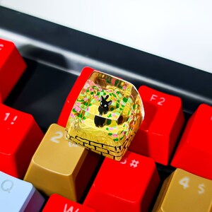 K&ouml;nnte beinhalten: Eine transparente Harztastenkappe mit gelbem und gr&uuml;nem Blumendesign und einer schwarzen Hasenfigur im Inneren. Die Tastenkappe liegt auf einer Tastatur mit roten und gelben Tasten.