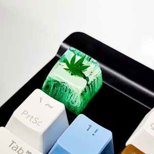 May include: A green and white keyboard keycap with a clear resin top featuring a green marijuana leaf. The keycap is labeled "Prtsc" and is on a black keyboard.