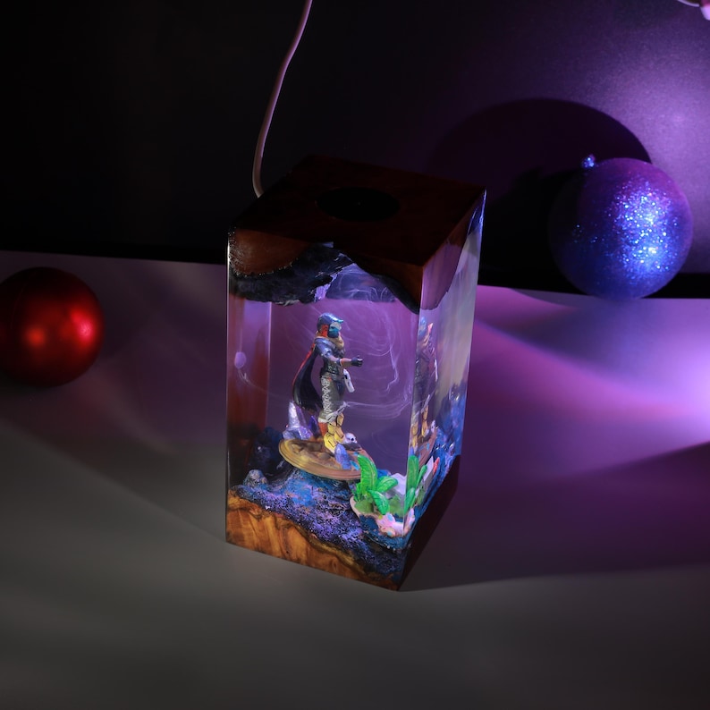 May include: A rectangular resin lamp featuring a dark wood top and base. Inside the clear resin, a miniature figure stands on a circular platform surrounded by small green plants and blue textured elements. The lamp emits a soft purple light.