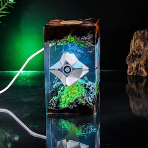 May include: A rectangular resin lamp featuring a white geometric Ghost character from Destiny suspended inside. The base and top are dark wood with jagged edges, while the interior glows with bright green light and blue accents, powered by a white cord.