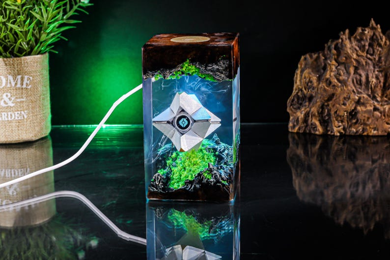 May include: A rectangular resin lamp featuring a white geometric Ghost character from Destiny suspended inside. The base and top are dark wood with jagged edges, while the interior glows with bright green light and blue accents, powered by a white cord.