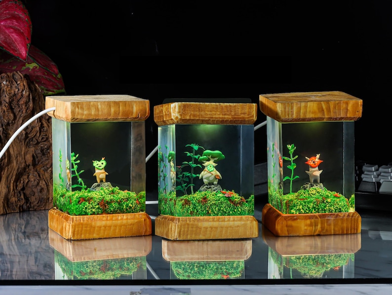 K&ouml;nnte beinhalten: Drei Lampen aus Holz mit klaren Glasw&uuml;rfeln, in denen sich jeweils eine kleine gr&uuml;ne, braune und wei&szlig;e Figur befindet. Die Figuren sind von gr&uuml;nem Moos und kleinen Pflanzen umgeben. Die Lampen sind von innen beleuchtet und strahlen ein warmes Licht aus.