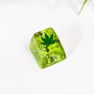 May include: A green and yellow resin keycap with a green marijuana leaf design.