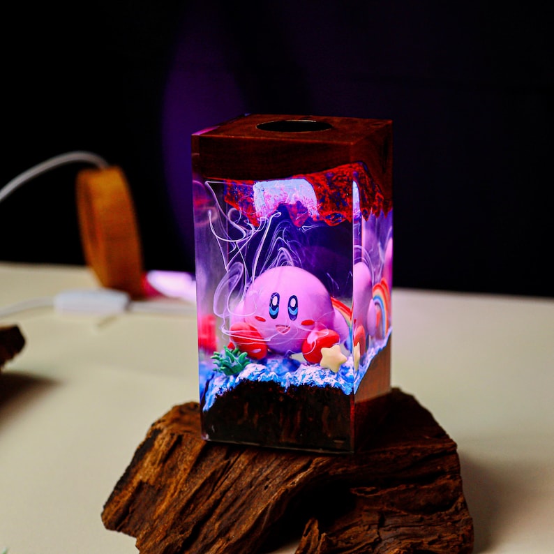 May include: A decorative wooden block with a pink Kirby figure inside a clear resin cube. The cube has a rainbow, stars, and other colorful elements. The wooden base is dark brown, and the resin glows with a purple and blue light.
