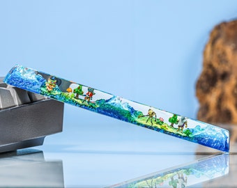 May include: A clear resin keyboard keycap with a miniature scene inside. The scene features a small forest with trees, a mushroom, and tiny figures. The resin is blue and green, and the figures are painted in bright colors.