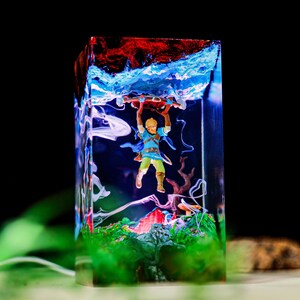 May include: A rectangular resin art piece featuring a miniature figure of a character from The Legend of Zelda. The figure is holding a wooden object, set against a backdrop of blue, red, and green colors, with a black background.