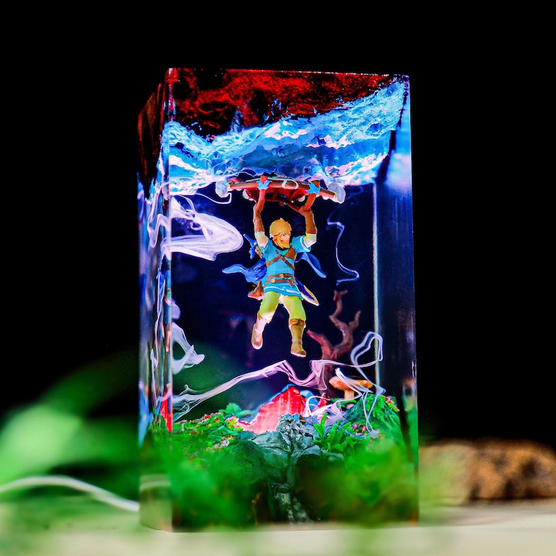 May include: A rectangular resin art piece featuring a miniature figure of a character from The Legend of Zelda. The figure is holding a wooden object, set against a backdrop of blue, red, and green colors, with a black background.
