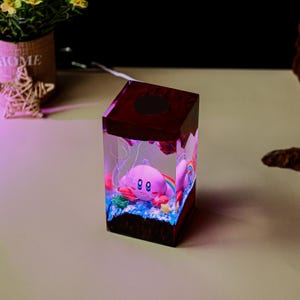 May include: A decorative resin cube featuring a pink cartoon character with a rainbow and blue accents. The cube has a dark brown top and base, with a light shining through the resin. The cube is on a light-colored surface.