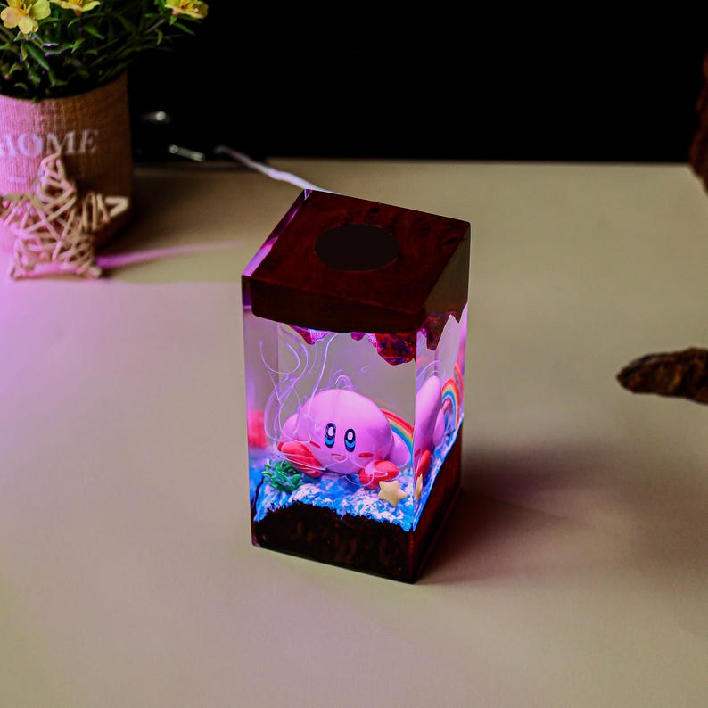 May include: A decorative resin cube featuring a pink cartoon character with a rainbow and blue accents. The cube has a dark brown top and base, with a light shining through the resin. The cube is on a light-colored surface.