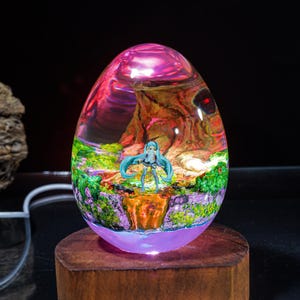 Puede incluir: Una l&aacute;mpara de vidrio transparente en forma de huevo con una escena colorida en su interior. La escena presenta a un personaje femenino de anime de pie en un bosque con un cielo rosa y morado. La l&aacute;mpara est&aacute; iluminada con una luz blanca c&aacute;lida.