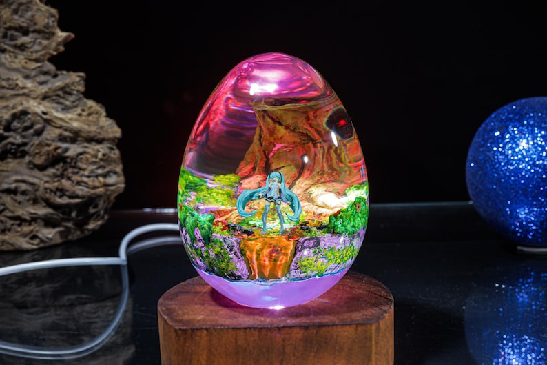 Puede incluir: Una l&aacute;mpara de vidrio transparente en forma de huevo con una escena colorida en su interior. La escena presenta a un personaje femenino de anime de pie en un bosque con un cielo rosa y morado. La l&aacute;mpara est&aacute; iluminada con una luz blanca c&aacute;lida.