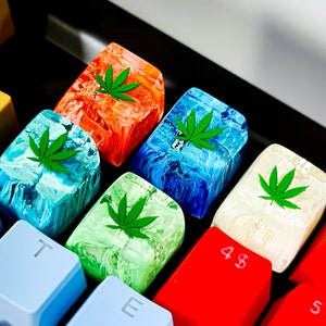 May include: Six colorful keyboard keycaps with a green marijuana leaf design. The keycaps are made of resin and have a swirled pattern. The keycaps are labeled with the letters "T", "E", "48", and "S".