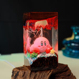 May include: A pink Kirby figurine inside a clear resin block with a wooden base. The resin has red and white accents, with a star and green details at the base. The figurine has black eyes and a smiling expression.