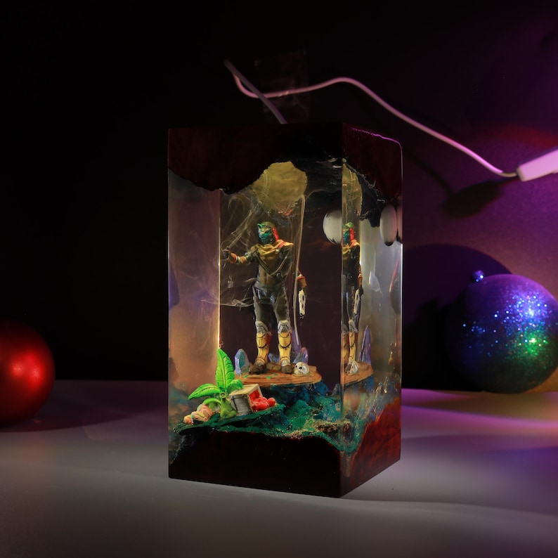 May include: A rectangular resin lamp featuring a miniature figure of a character with a green mask and yellow armor standing on a base with a small green plant and a crate. The clear resin block is encased in dark wood at the top and bottom.