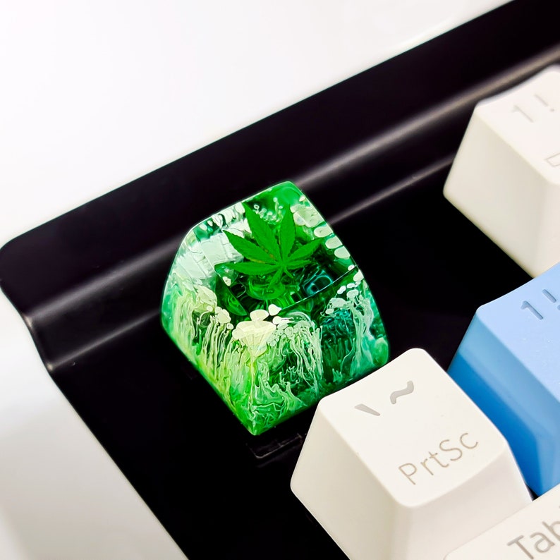 May include: A green and white resin keycap with a green leaf design. The keycap is on a black keyboard.