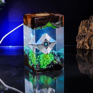 May include: A rectangular resin lamp featuring a dark wood top and base with a clear center section. Inside the resin, a white geometric robot-like figure with a central blue eye is suspended above a vibrant green moss-like texture.