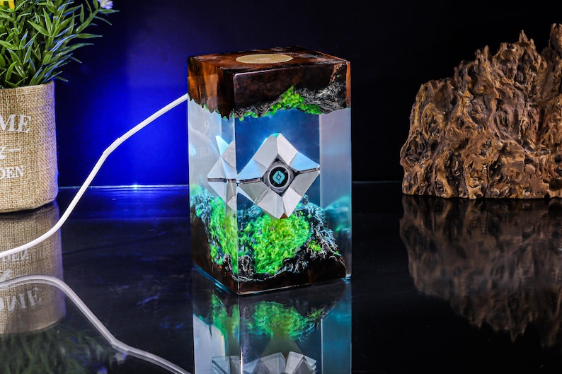May include: A rectangular resin lamp featuring a dark wood top and base with a clear center section. Inside the resin, a white geometric robot-like figure with a central blue eye is suspended above a vibrant green moss-like texture.
