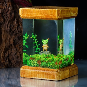 K&ouml;nnte beinhalten: Eine kleine Holzkiste mit einem transparenten Harzeinsatz, der zwei kleine gr&uuml;ne Figuren und gr&uuml;ne Pflanzen enth&auml;lt. Die Kiste ist aus Holz mit einer nat&uuml;rlichen Oberfl&auml;che.