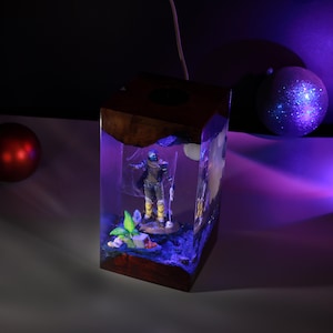May include: A rectangular lamp made of dark wood and clear resin, featuring an internal diorama of a standing figure in a desert-like setting. The scene includes a small green plant, a skull, and a glowing blue light effect inside the resin.
