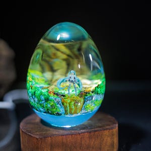 Puede incluir: Un orbe de vidrio transparente con un paisaje verde y azul en su interior. Una peque&ntilde;a figura de una mujer con el pelo largo azul se encuentra en el centro del paisaje.