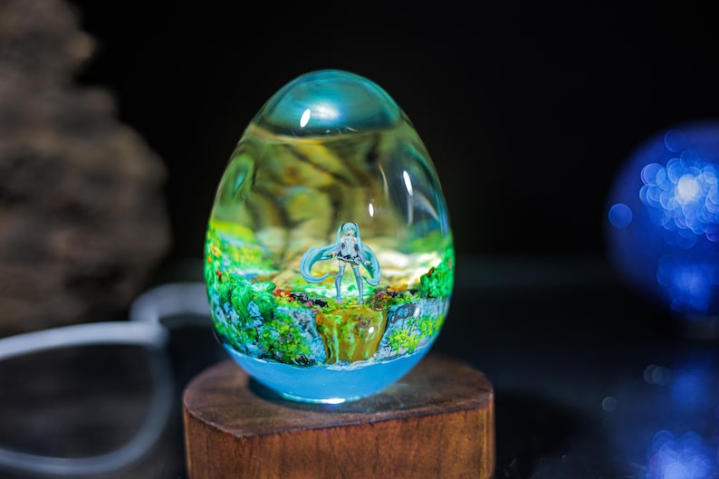 Puede incluir: Un orbe de vidrio transparente con un paisaje verde y azul en su interior. Una peque&ntilde;a figura de una mujer con el pelo largo azul se encuentra en el centro del paisaje.