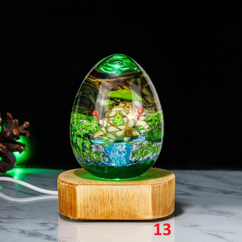 K&ouml;nnte beinhalten: Eine gr&uuml;ne Glaskugel mit einer Waldlandschaft im Inneren, in der sich ein kleines gr&uuml;nes Wesen unter einem Pilz befindet. Die Kugel steht auf einem Holzsockel.