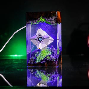 May include: A rectangular resin lamp featuring a white geometric Ghost character from Destiny suspended inside. The clear resin block contains dark rocky textures, vibrant green moss-like elements, and purple internal lighting.