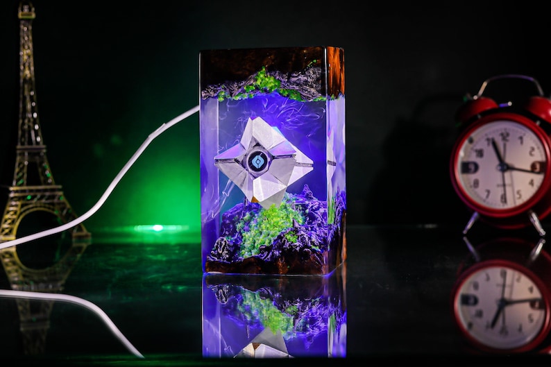 May include: A rectangular resin lamp featuring a white geometric Ghost character from Destiny suspended inside. The clear resin block contains dark rocky textures, vibrant green moss-like elements, and purple internal lighting.