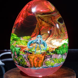 Puede incluir: Una esfera de vidrio transparente con un paisaje rojo, naranja y verde en su interior. Una peque&ntilde;a figura azul y blanca de una mujer con el pelo largo azul se encuentra en el centro de la esfera.