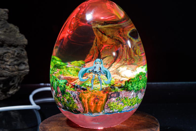 Puede incluir: Una esfera de vidrio transparente con un paisaje rojo, naranja y verde en su interior. Una peque&ntilde;a figura azul y blanca de una mujer con el pelo largo azul se encuentra en el centro de la esfera.
